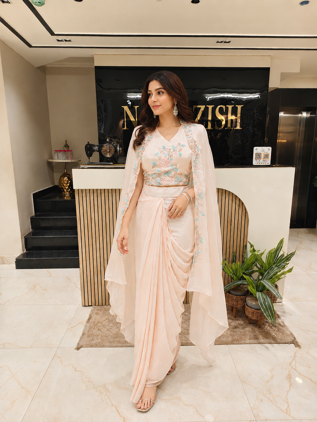 Woman in a light pink saree with a floral blouse standing in a modern interior setting.