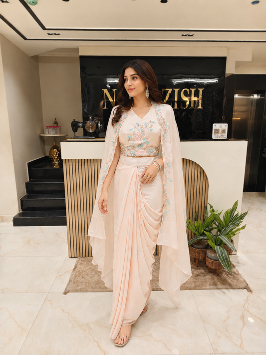 Woman in a light pink saree with a floral blouse standing in a modern interior setting.